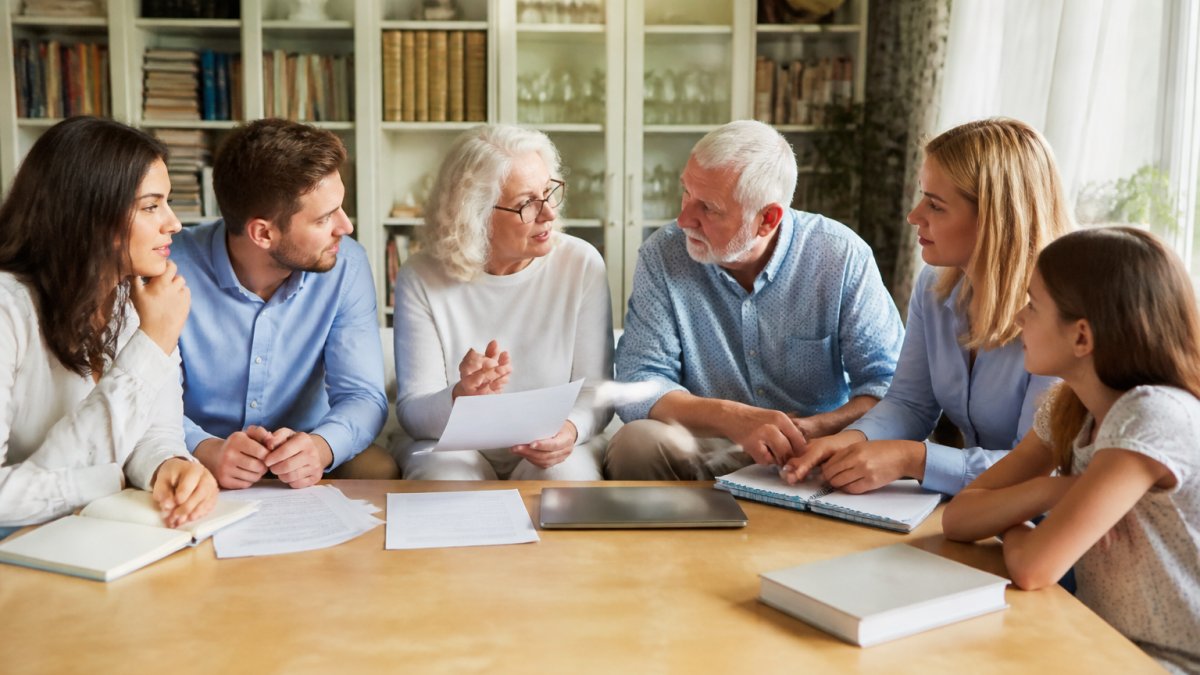 A multi-generational family seated around a large wooden table in a bright home office, with notebooks and a laptop open, discussing financial plans together