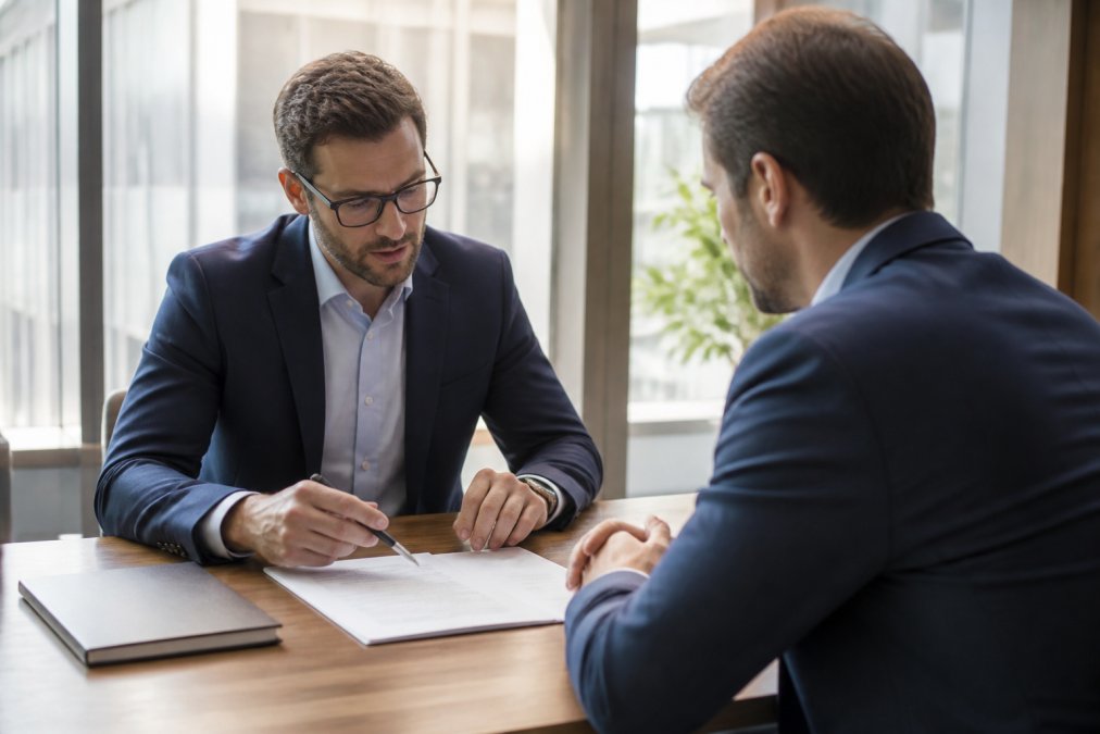 Zwei Geschäftsleute sitzen sich in einem modernen Büro gegenüber. Eine Person hält ein Dokument oder einen Stift, während beide auf einen Vertrag oder eine Checkliste auf dem Tisch blicken. Vertrauensvoll und professionell. Natürliches Licht durch große Fenster.