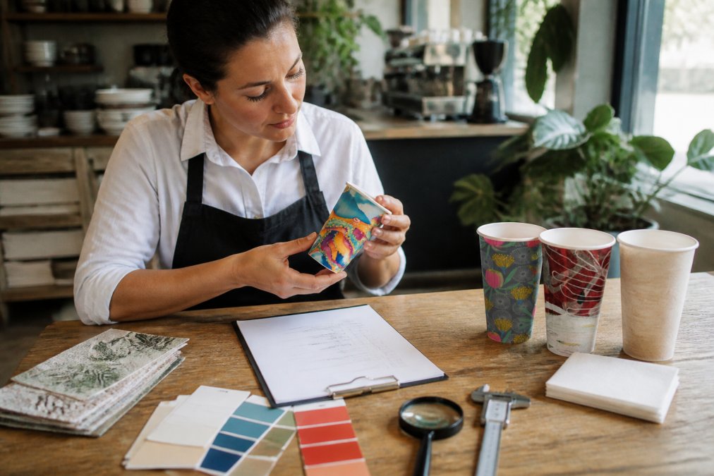 Cafe manager reviewing branded compostable cup samples and napkin designs at a wooden table with a printed checklist, colour swatches, and quality inspection tools under natural window lighting