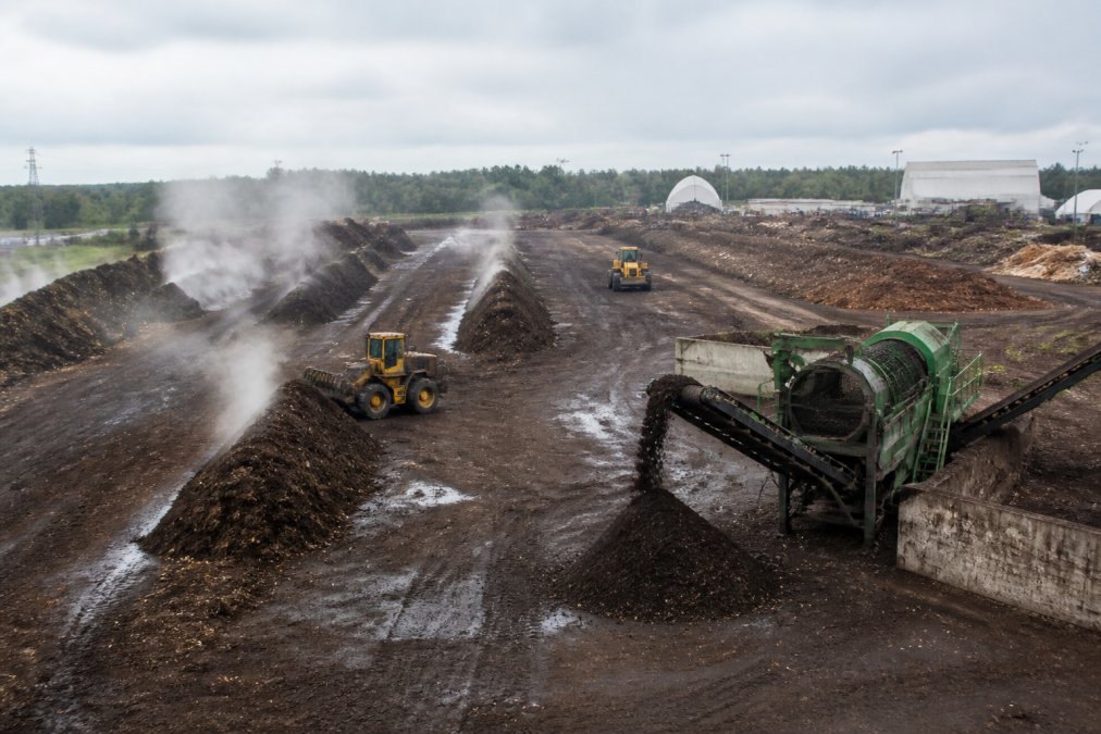 Large industrial composting facility with rows of composting windrows being turned by heavy machinery, steam rising from organic material decomposition, metal screening equipment sorting finished compost under grey overcast sky