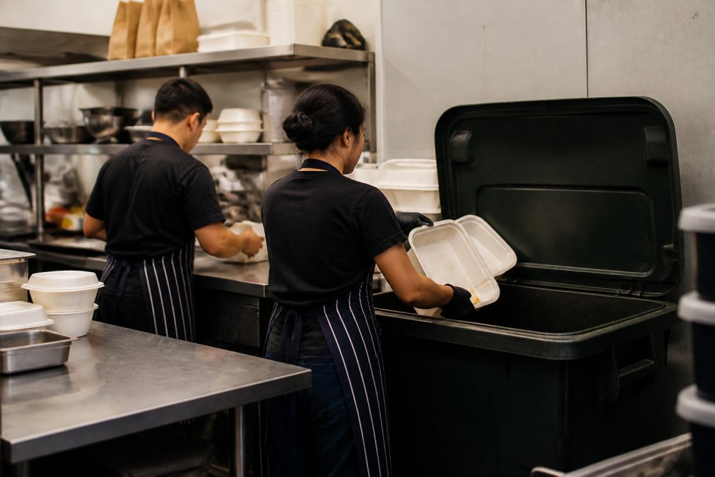 Restaurant staff in aprons placing used white compostable takeaway containers into a large collection bin marked for composting, demonstrating the disposal process in a commercial kitchen