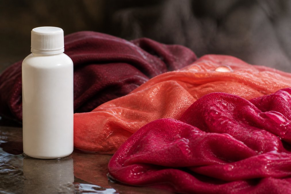 Una bottiglia di fissativo professionale accanto a tessuti colorati appena tinti, con gocce d'acqua e vapor visibili sulla superficie