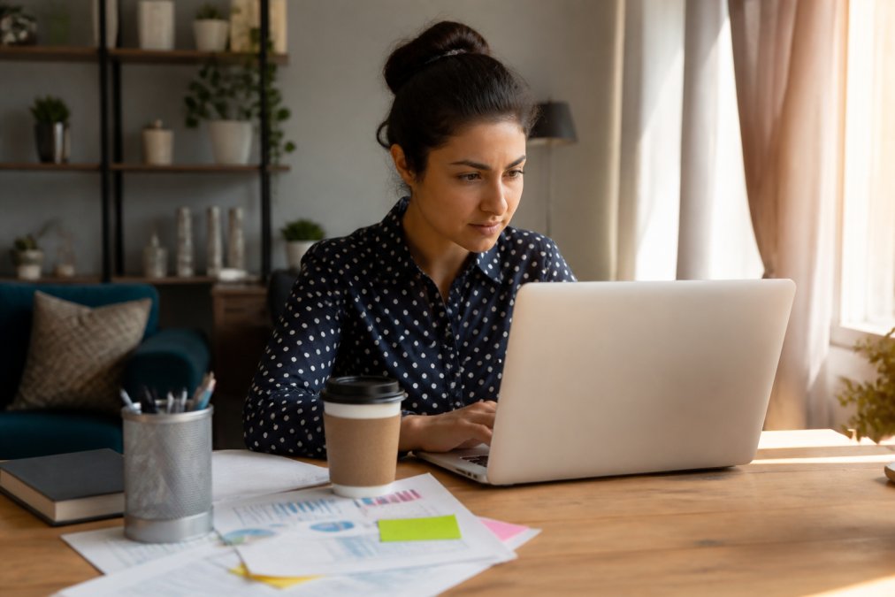 Freelancer working at home office desk with laptop, coffee, and business documents spread across the surface, natural morning light streaming through a window, looking focused on project work