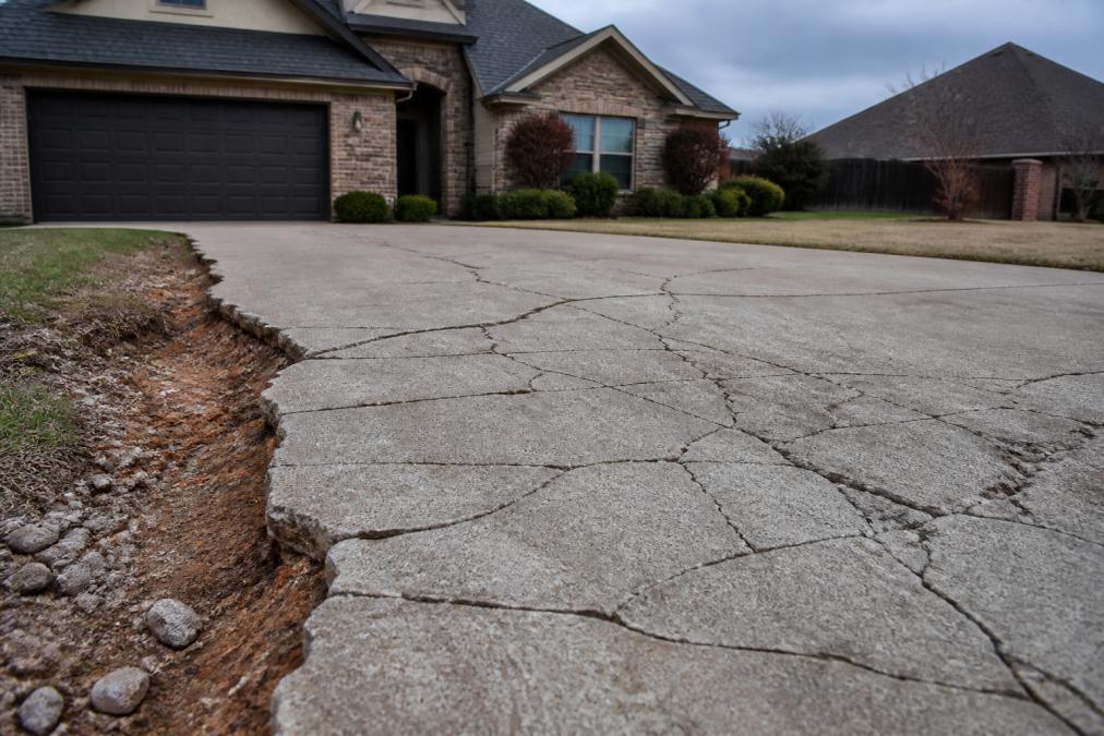 A cracked concrete driveway in a residential neighborhood with visible spider-web fractures, clay soil exposed at the edge, and a house in the background under overcast Oklahoma skies