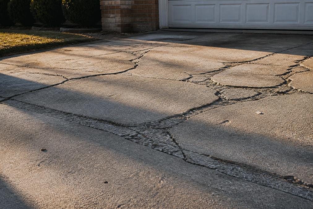 Close-up photo of a residential concrete driveway with deep, wide cracks and spalling surface, uneven slabs visible, harsh afternoon sunlight casting long shadows across the damaged concrete