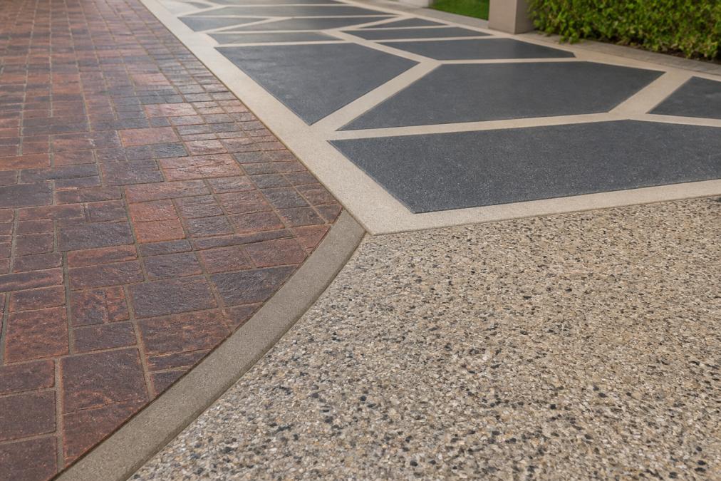 Close-up detail of a residential driveway showing multiple decorative concrete finishes, a stamped pattern section with brick-like texture, an adjacent stencilled geometric design in contrasting colours, and a smooth honed concrete section with polished aggregate exposed