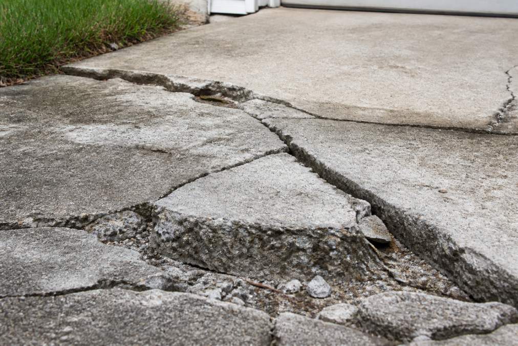 Close-up of severely cracked and spalling concrete driveway surface with deep fissures, deteriorated edges, and visible damage exposure in natural daylight