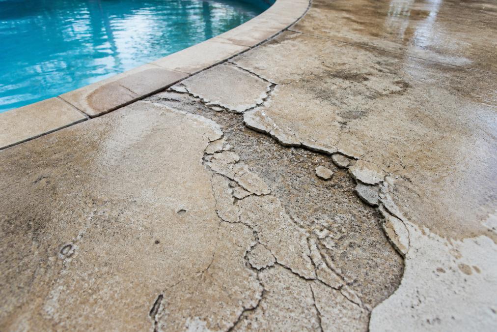 Close-up of a cracked and deteriorating pool deck concrete surface with visible spalling, flaking edges, and water-stained areas showing extent of damage before professional repair