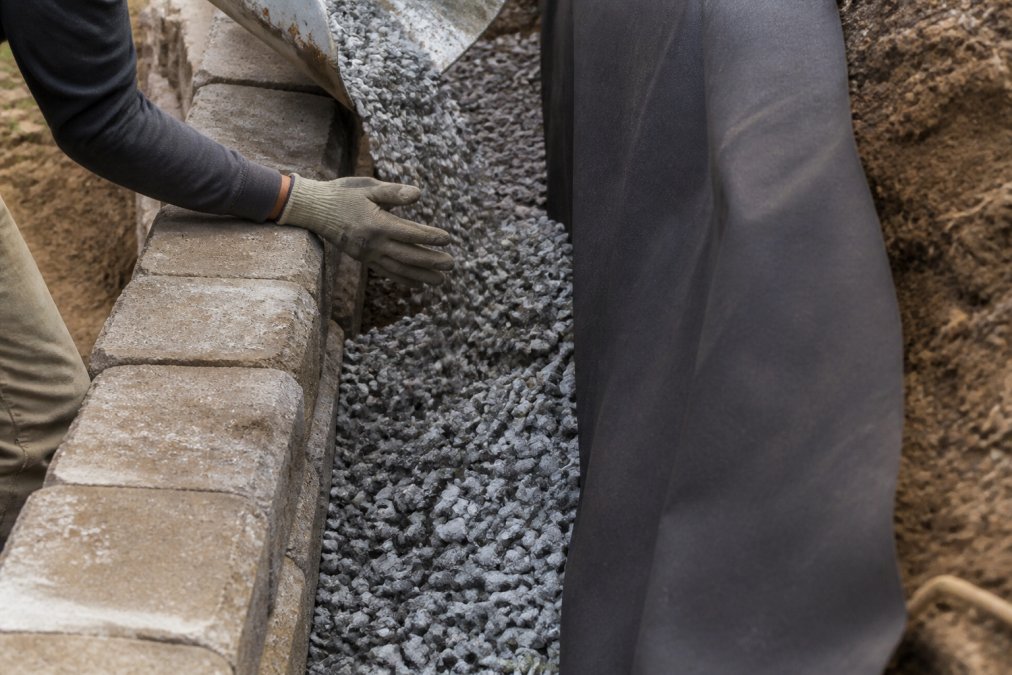 Close-up of crushed angular gravel being poured behind a concrete block retaining wall, geotextile fabric visible against the soil, worker's gloved hands guiding the stone