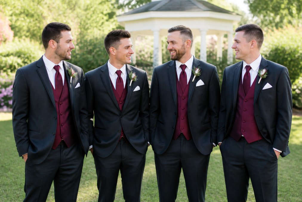 A group of four groomsmen in coordinated charcoal formal suits with matching burgundy waistcoats, neckties and white pocket squares, standing outdoors at a garden wedding venue with soft natural afternoon light, looking polished and relaxed