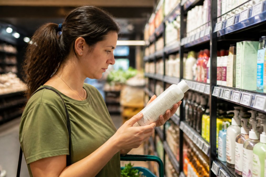 A shopper in a bright Australian retail store carefully examining the back of a product package, holding it close to read small certification text, surrounded by shelves of packaged goods in warm overhead lighting