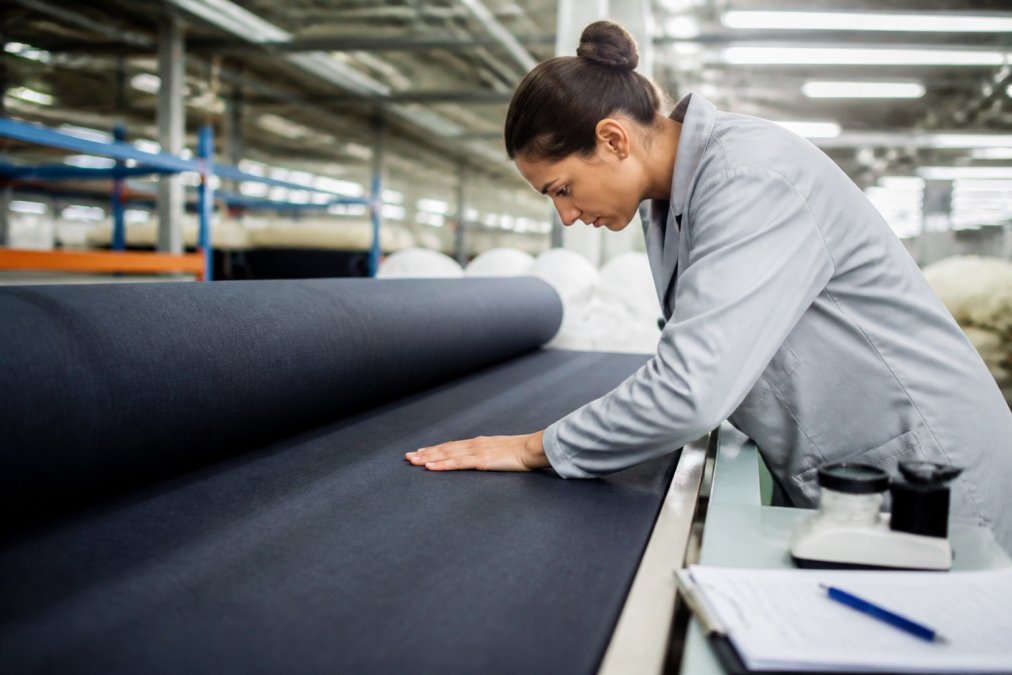 Quality control specialist inspecting finished fabric under bright overhead lighting, checking for texture consistency and color uniformity on a bolt of material, with testing equipment and documentation visible