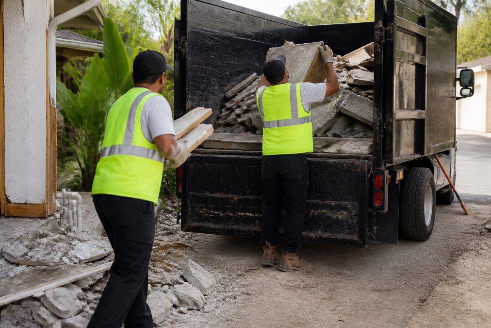 Professional junk removal crew in safety vests loading construction debris including drywall, lumber, and concrete pieces into a large truck bed on a residential job site in daylight