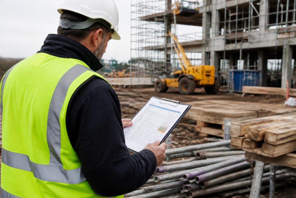 A site supervisor in high-visibility clothing and hard hat reviewing a printed risk assessment on a clipboard while standing on a partially built construction site, with scaffolding and plant machinery visible in the background
