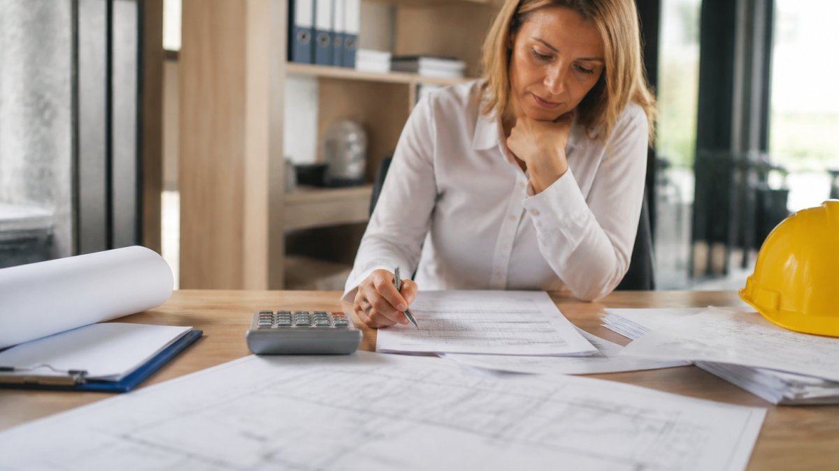Office manager reviewing construction project budgets and cost spreadsheets on a computer screen with blueprints, cost documents, and calculator visible on desk