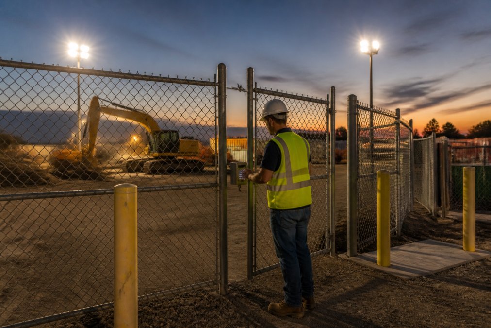 A construction site at dusk with chain-link fencing, motion-activated floodlights illuminating heavy equipment, and a locked gate with access control keypad