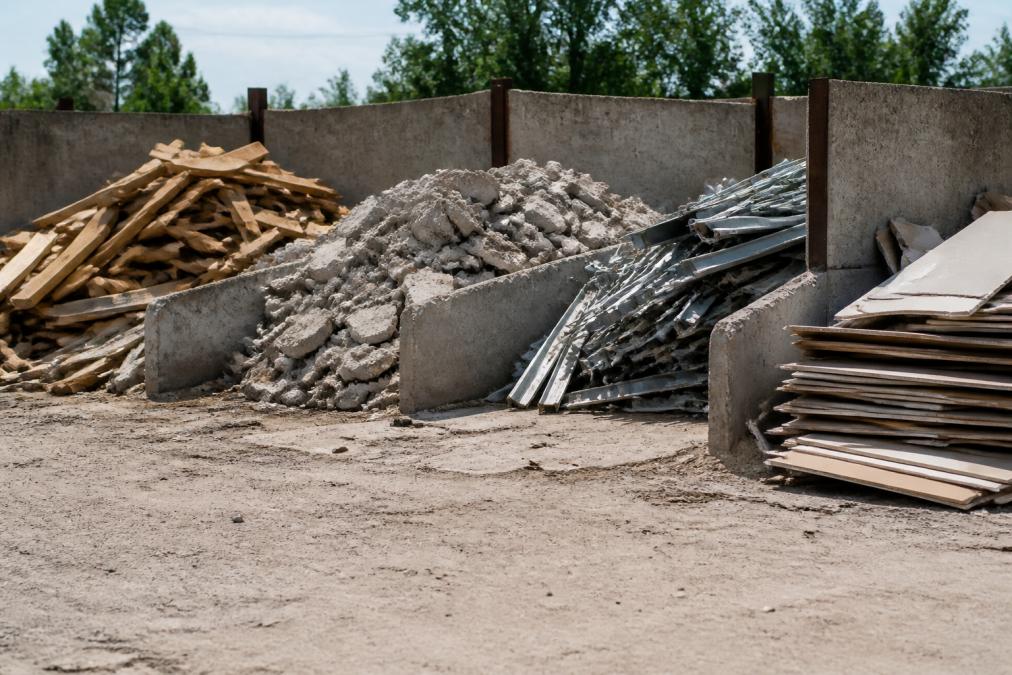 Construction site with organized piles of sorted debris including stacked lumber, concrete chunks, metal scraps, and separated drywall sheets in clearly marked sections with warning signage