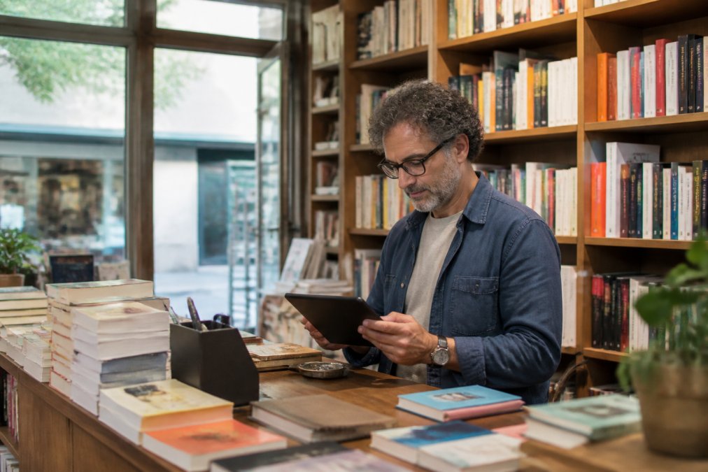 Libraio indipendente seduto al bancone di una libreria accogliente che consulta dati di vendita su un tablet, con scaffali di libri colorati sullo sfondo e luce naturale che filtra da una vetrina