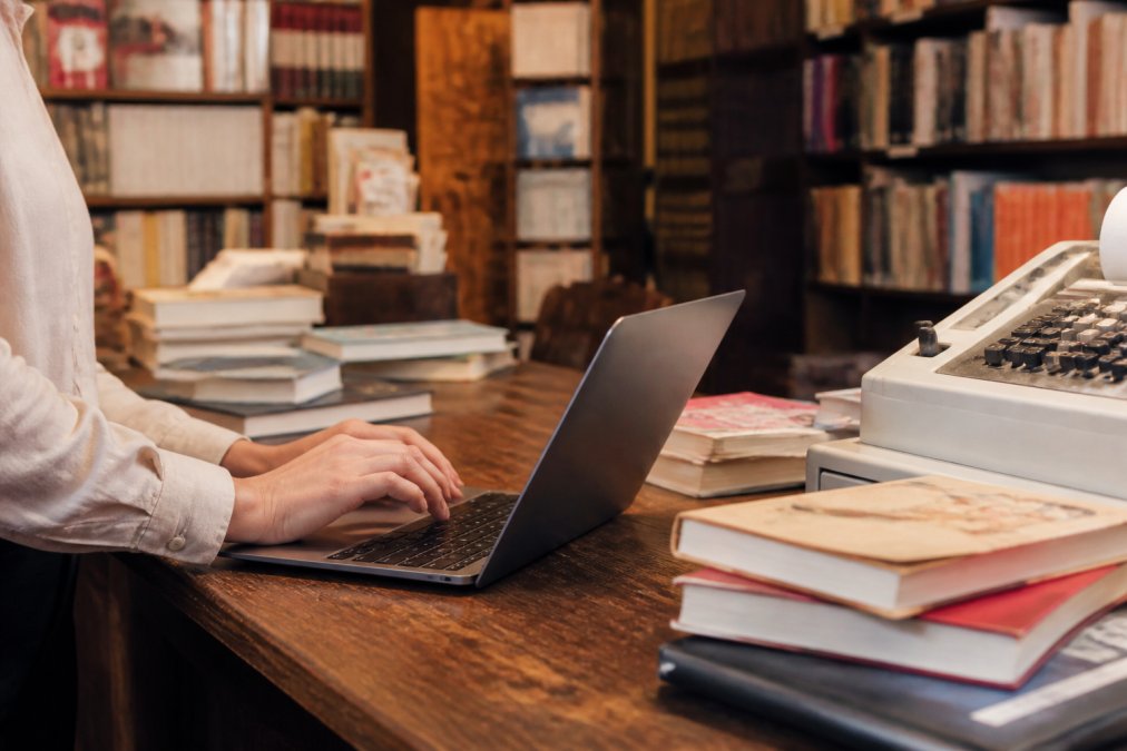 Mani di una persona che digitano su un laptop aperto sul bancone di una libreria indipendente accogliente, con pile di libri e un piccolo registratore di cassa visibili sul tavolo, illuminazione calda e soffusa