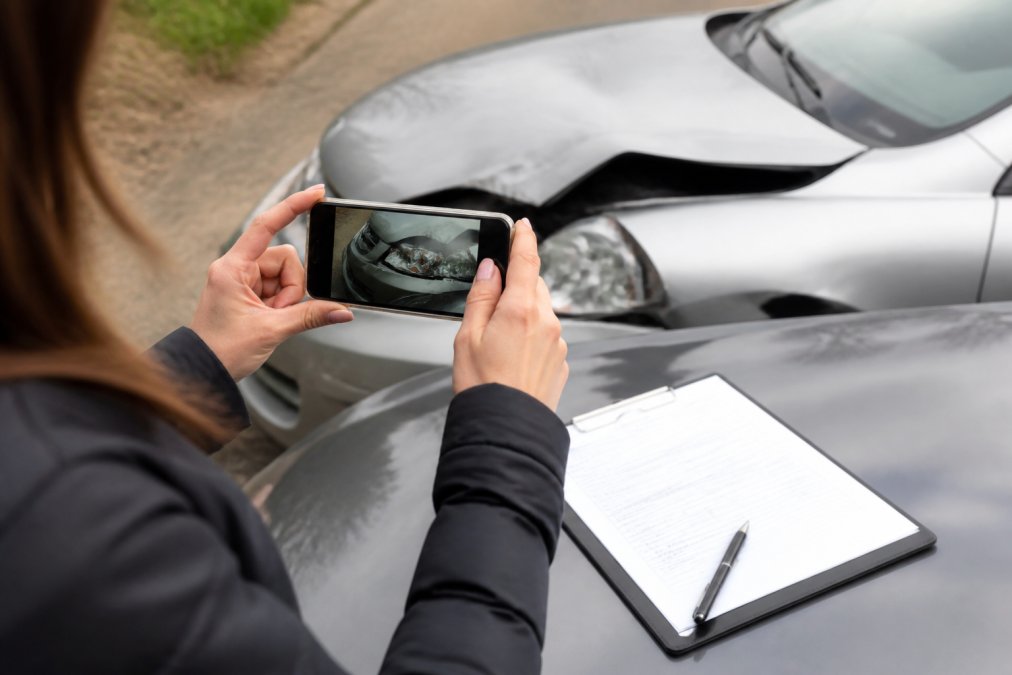 Person carefully photographing vehicle damage at accident scene with smartphone, accident report form and clipboard visible on hood, overcast daylight
