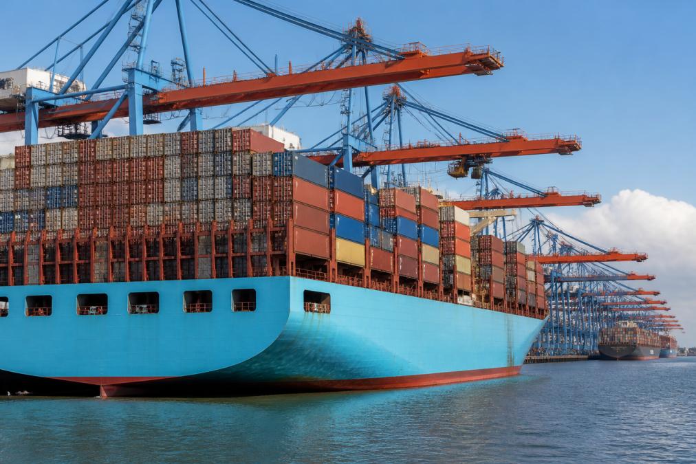 A massive container ship docked at a busy port terminal with tall stacks of colorful shipping containers and large gantry cranes loading cargo under a clear sky