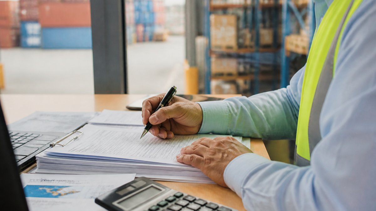 Logistics professional reviewing container tracking details on computer screen at desk with shipping documents and warehouse background