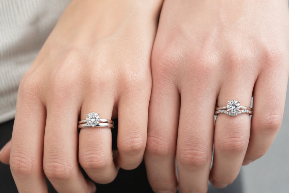 Close-up of a hand wearing a straight wedding band next to an engagement ring, showing a visible gap, and another hand with a contour band sitting flush against the ring