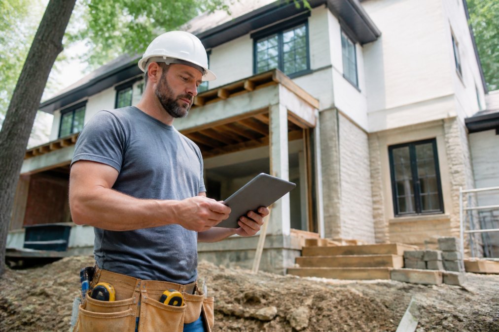 Contractor reviewing project plans on a tablet while standing next to a residential home under renovation, natural daylight
