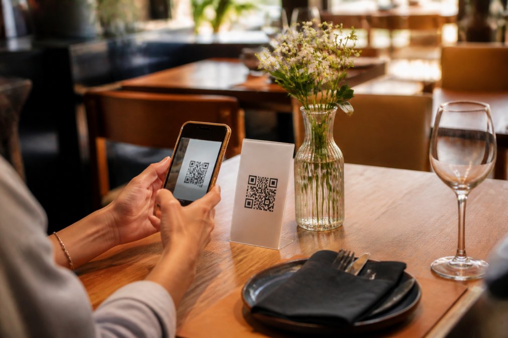 Comensal en una mesa de restaurante moderno escaneando un código QR con su smartphone mientras sostiene el dispositivo sobre la mesa, con el menú digital visible en la pantalla brillante