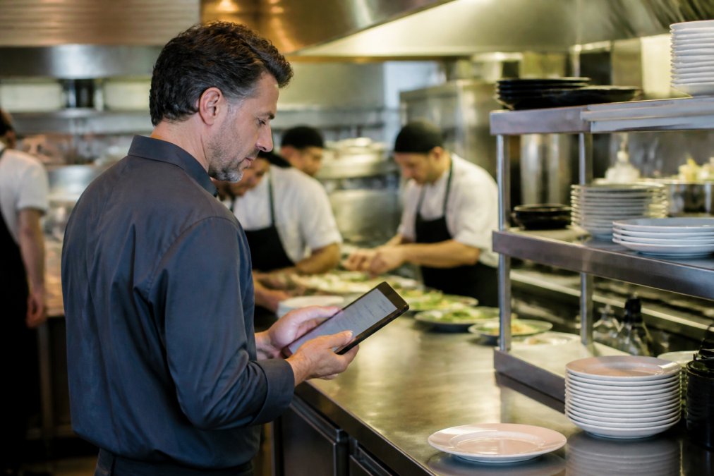 Gerente de restaurante usando una tableta para actualizar precios y disponibilidad de platos mientras trabaja en la cocina, con el panel de control visible en la pantalla
