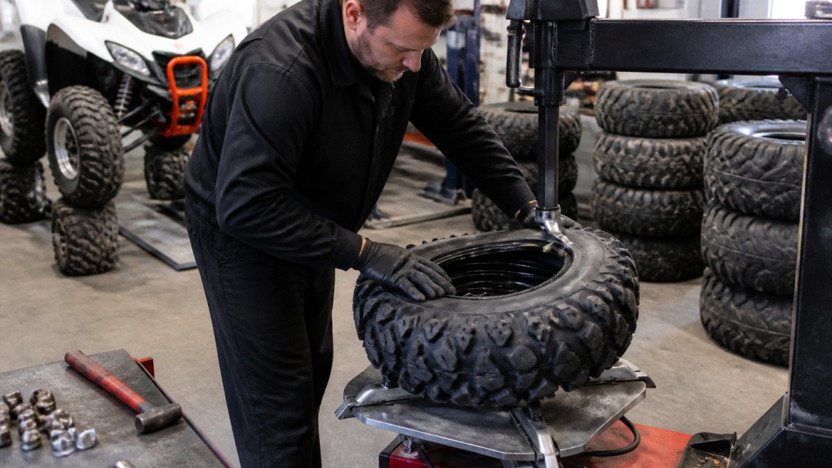 A technician in a service bay mounting a new knobby ATV tire onto a rim, with a stack of tires and tools visible in the background