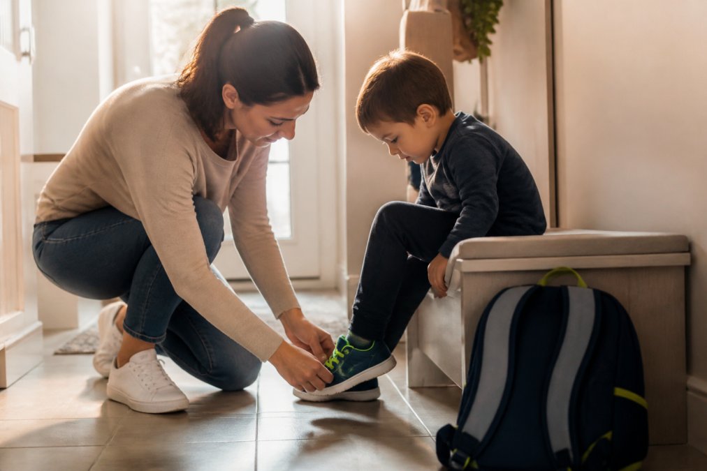 A parent kneeling on a hallway floor helping a young child slip on a sneaker with bright elastic laces, school backpack visible nearby, warm morning light