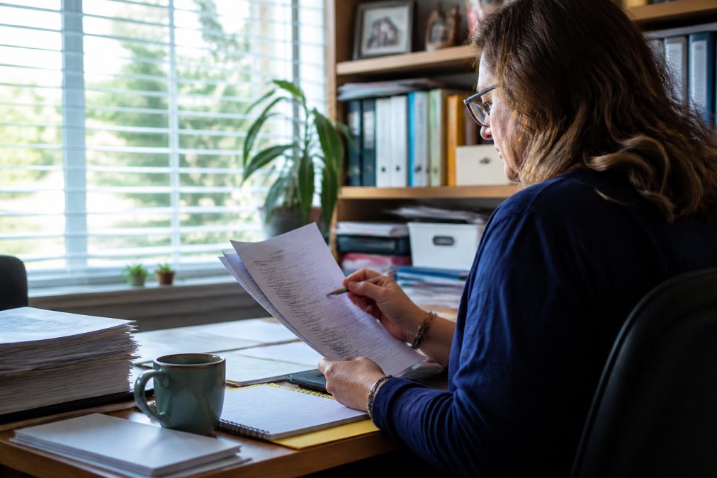 Educator reviewing student assessments on computer monitor in focused workspace, with coffee and organized desk materials, natural afternoon light from window