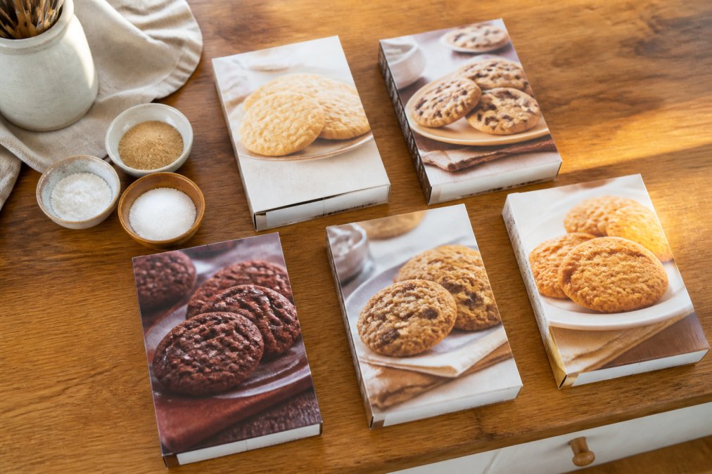 Overhead flat-lay of several labeled cookie mix boxes arranged on a warm wooden kitchen counter alongside small bowls of organic cane sugar, sea salt, and baking soda in natural morning light
