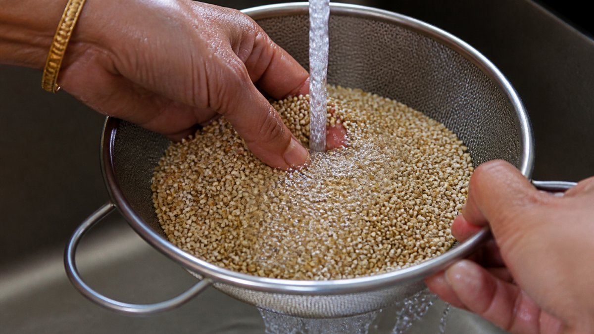 Close-up of hands rinsing unpolished millet in a fine-mesh strainer under running water, showing the small grains and water draining through the mesh