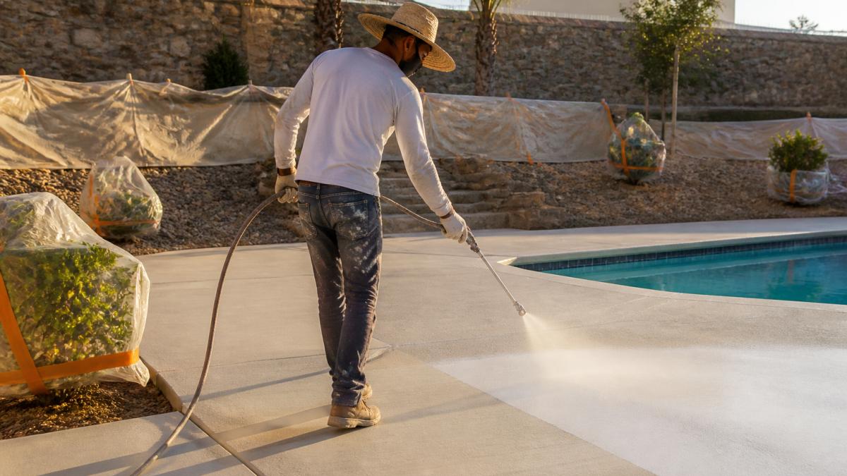 Professional contractor in safety gear applying Cool Deck coating to concrete pool deck using spray equipment, with protective coverings visible around landscaping