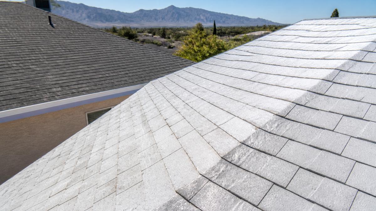 Close-up of reflective cool roof shingles installed on a residential home with bright sunlight reflecting off the surface, showing the light color contrast against a traditional dark roof in the background