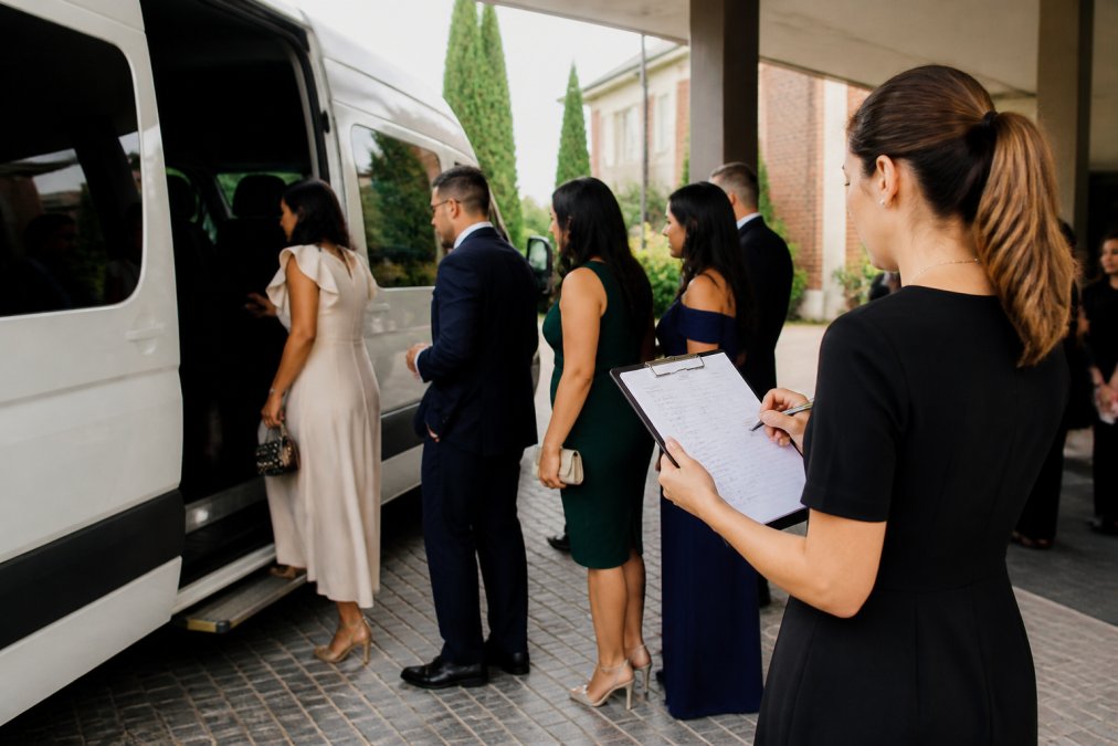 A wedding guest coordinator holding a clipboard and checking names off a list while guests board a white minibus outside a hotel entrance