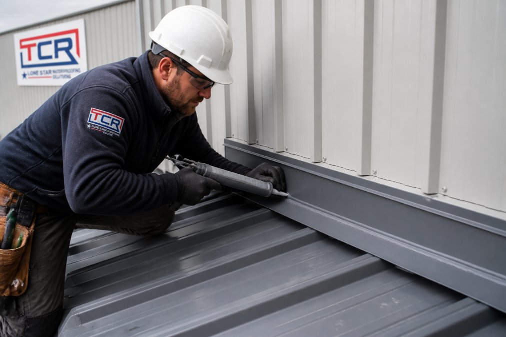 Contractor applying sealant and flashing while coordinating roof and wall panel repairs on an industrial building.