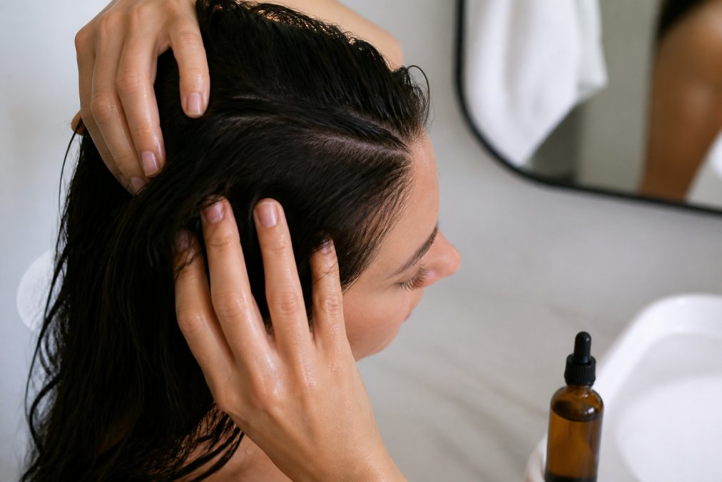 Close-up of hands applying copper peptide serum to scalp with gentle massage motion, showing proper fingertip application technique with a bottle of peptide serum nearby on a light bathroom counter