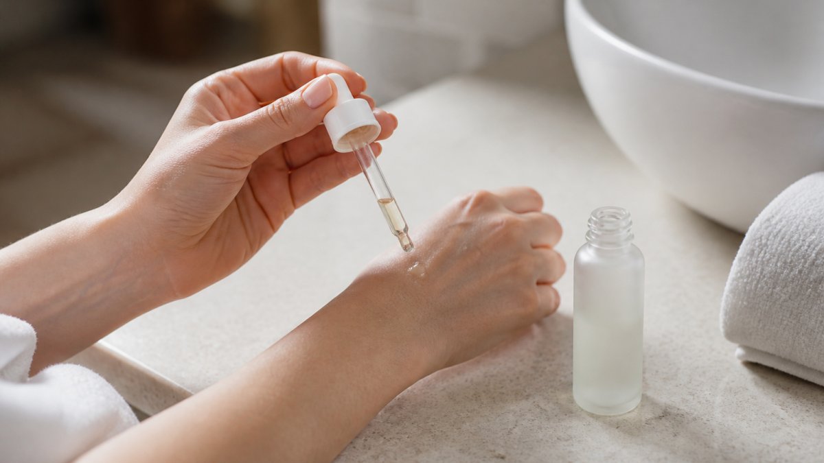 Close-up of a woman's hands applying a drop of lightweight serum to her forearm, with a minimalist skincare bottle and soft natural light on a bathroom counter