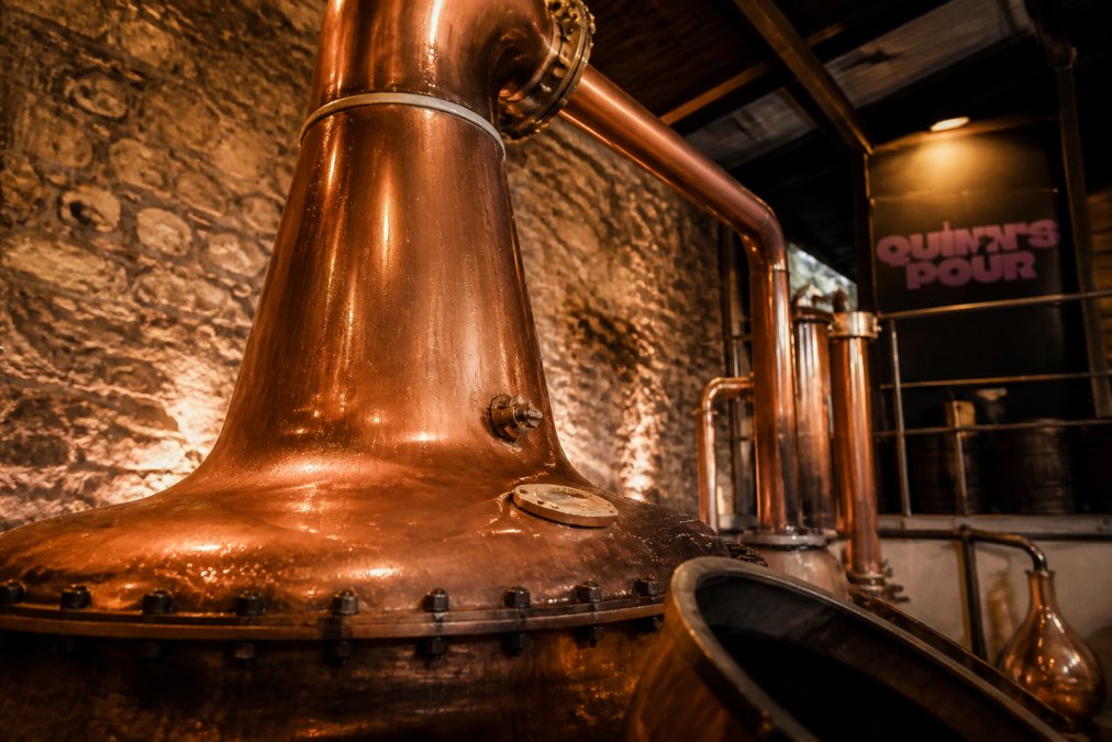 Close-up of a traditional copper pot still in a small distillery, with condensation visible on the curved copper surface and warm ambient lighting