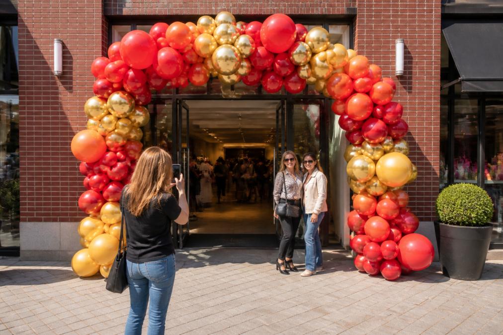 A wide, colorful balloon arch framing the entrance of a brick retail storefront at a grand opening, with branded balloons in coordinated red and gold brand colors, a lush balloon garland cascading down both sides, and customers pausing to take photos in bright afternoon sunlight