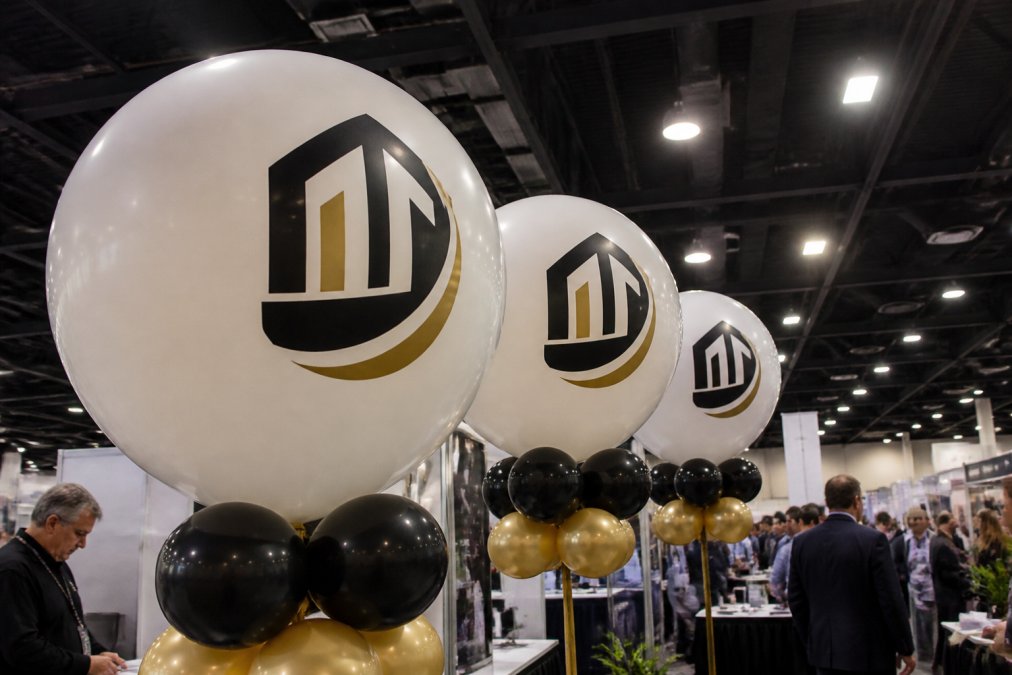 Close-up of large round white latex balloons printed with a bold black company logo and gold accent brand colors, arranged in a cluster display at a corporate event trade show booth, with soft overhead lighting and guests visible in the background