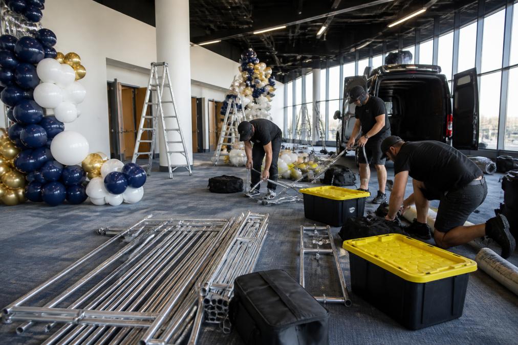 Team of professional decorators efficiently breaking down and packing away balloon installation equipment from a high-ceiling corporate venue, carefully folding and storing frameworks, coiling cables, and organizing supplies into transport vehicles