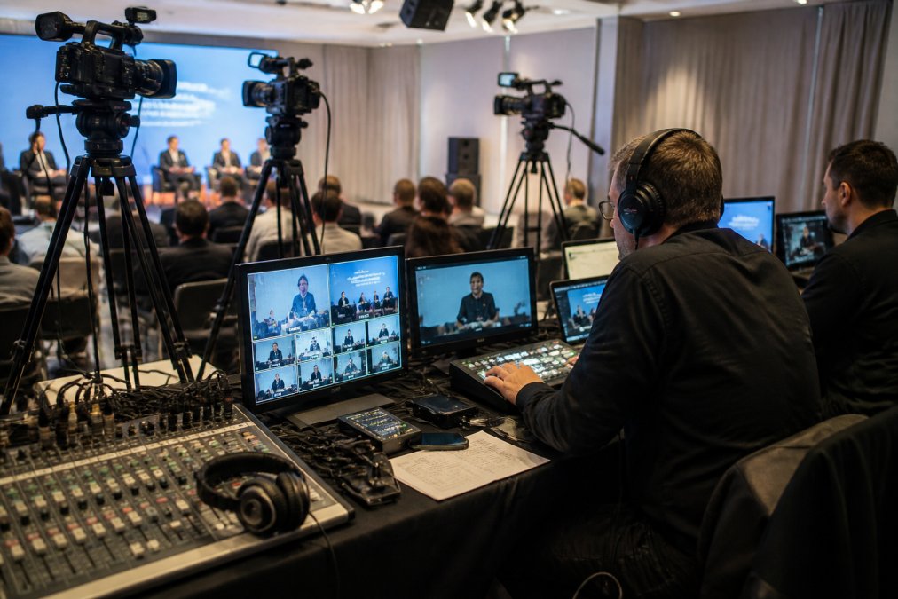 Professional streaming setup showing multiple 4K cameras mounted on tripods, audio mixing console with faders and monitoring headphones, video switcher with multiple input screens, and technicians in corporate event space monitoring live broadcast on control room displays with real-time analytics visible