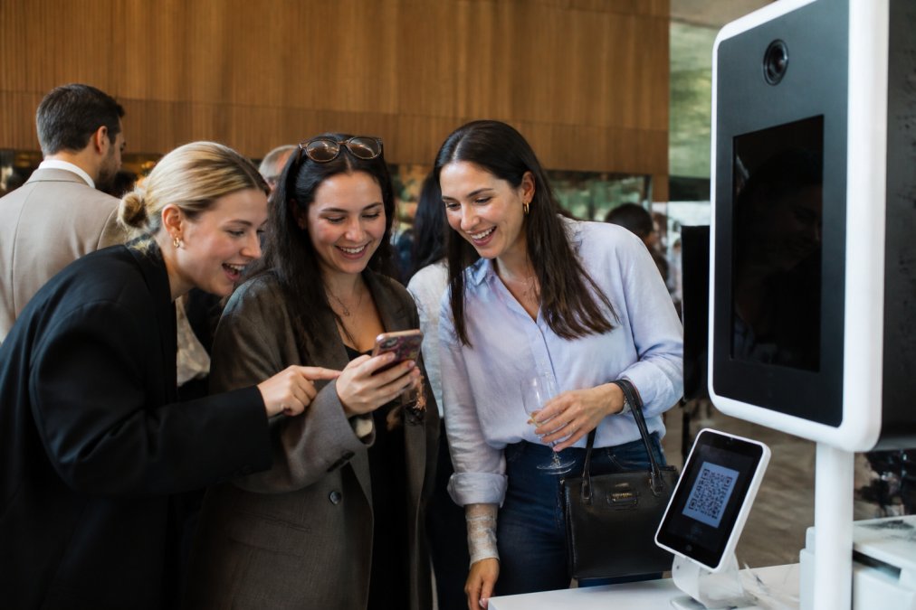 Professional demonstrating corporate event photo booth branding tips with attendees smiling while viewing their photo booth image on a smartphone, with a branded photo booth visible in the background and a QR code displayed on a tablet