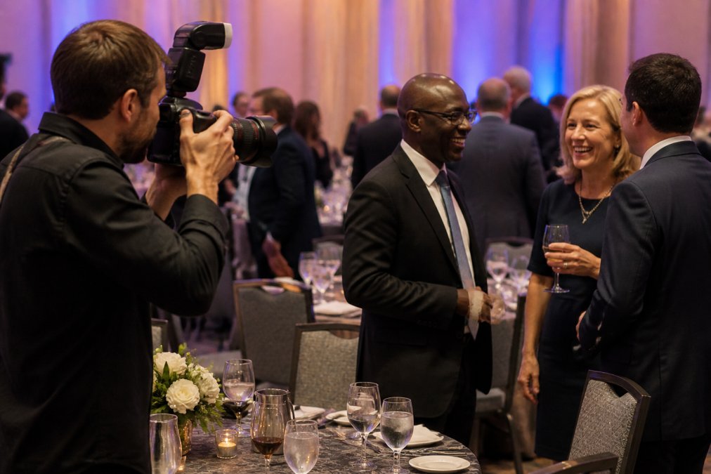 Professional photographer capturing candid moments between corporate attendees at an elegant gala, with table settings and networking conversations visible in soft ambient lighting