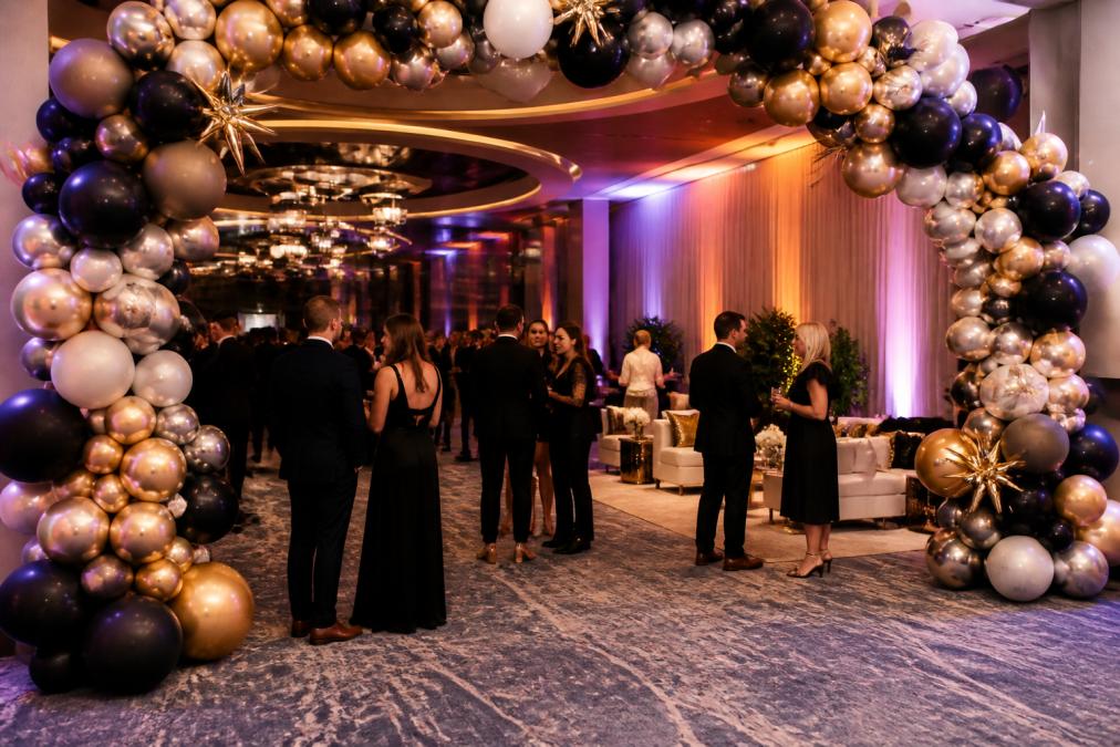 An elegant corporate gala with custom balloon arches framing the entrance, ambient uplighting casting soft colors across the venue, and professionally dressed guests mingling near a branded lounge area with curated furniture and floral centerpieces