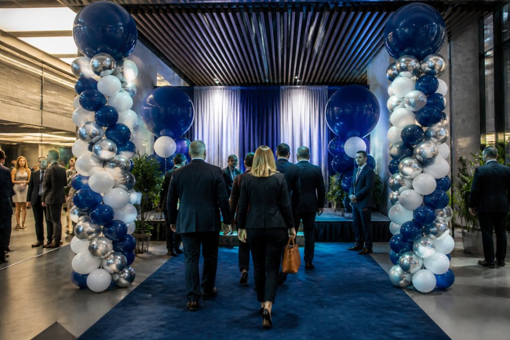 A grand opening event entrance featuring custom branded balloon installations in corporate colors, tall columns flanking a professional stage area, with ambient lighting highlighting the balloon work and guests in business attire walking through the branded entrance